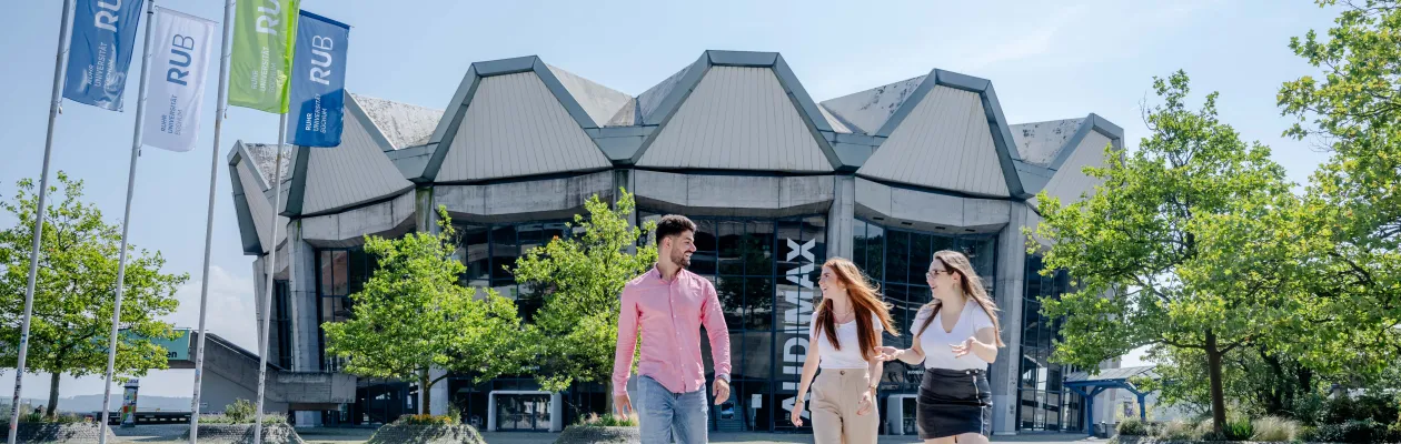 Foto: Drei Personen, ein Mann und zwei Frauen, gehen über den Forumsplatz vor dem Audimax. Im Hintergrund sieht man das komplette Audimax mit der gleichlautenden Aufschrift. Links im Bild befinden sich mehrere Fahnen mit dem RUB-Logo. Die drei Personen schauen sich an, reden und lachen.