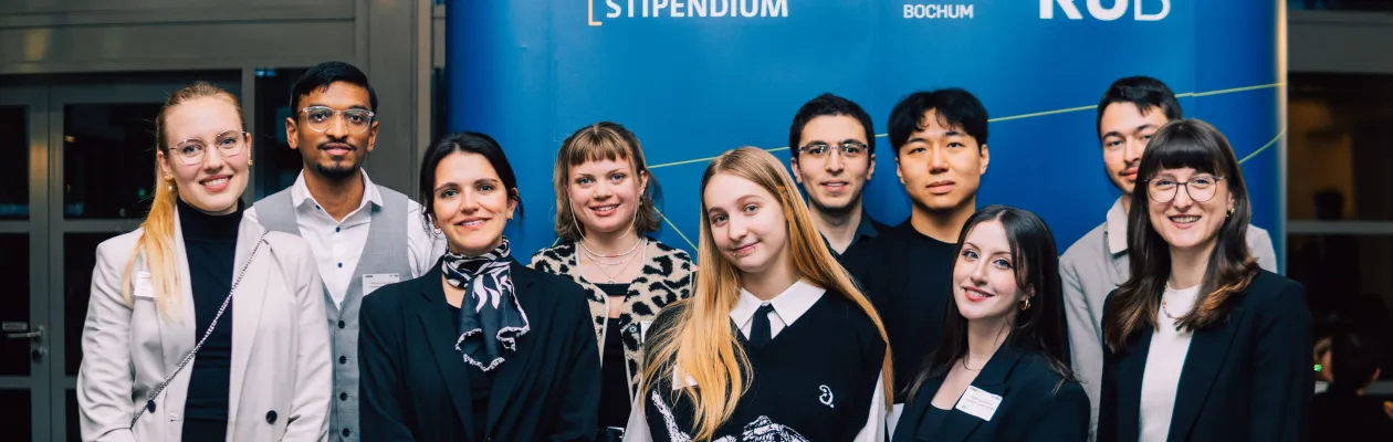 Foto: Mehrere Personen stehen lächelnd vor einem blauen Aufsteller. Auf dem Aufsteller befinden sich die Logos vom Deutschlandstipendium und von der Ruhr-Universität Bochum.