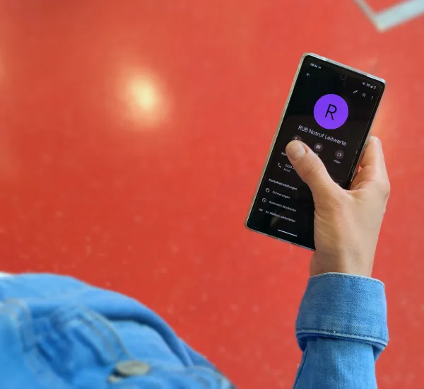 Hand hält Handy, darauf zu sehen der Kontakt RUB Leitwarte Notfall