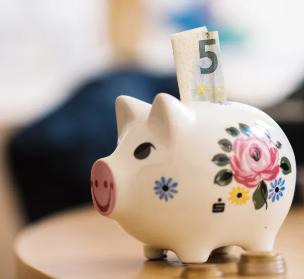 Foto: Ein Sparschwein mit Blumenmuster. Oben guckt ein 5 Euro-Schrein raus.