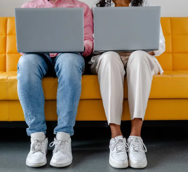 Foto: Zwei Personen sitzen nebeneinander. Beide haben einen aufgeklappten Laptop auf dem Schoß. Man sieht nur Füße und Beine der Personen sowie die Rückseite der Laptops.